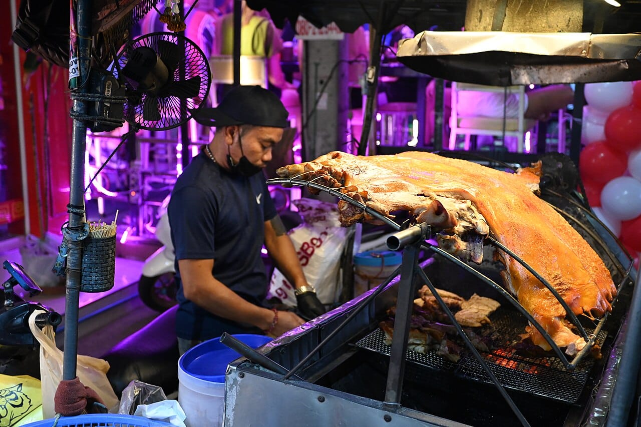 DZ3 0976 A street food vendor grilling a whole roasted pig over charcoal at a lively night market stall in Pattaya Thailand