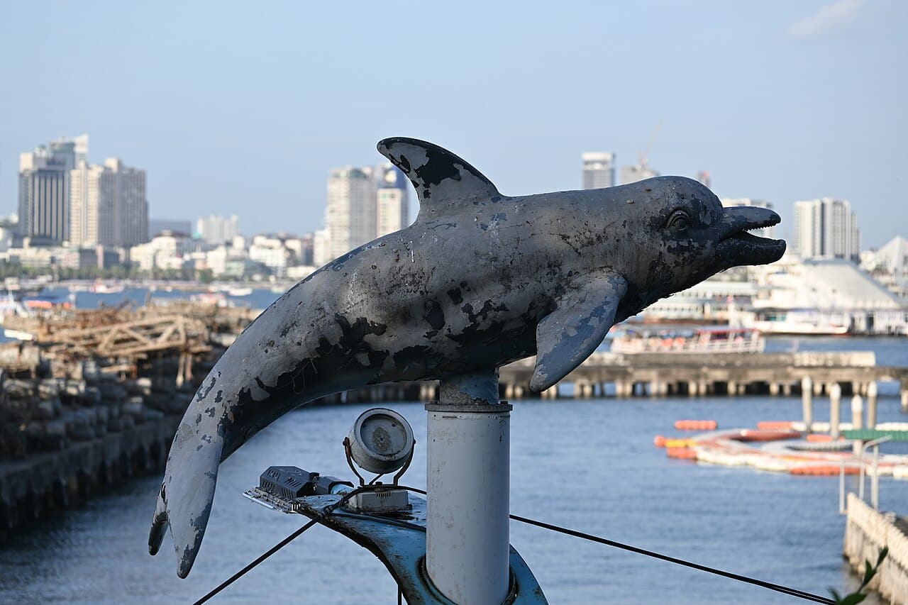DFC 1623 Weathered dolphin sculpture mounted on a harbor railing with city skyline and boats in the background